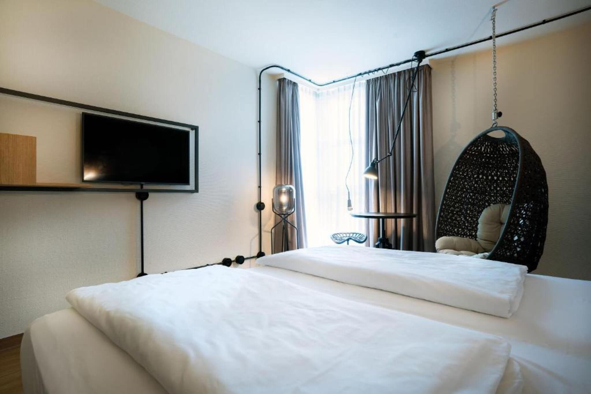 A hotel room with a bed, a television, a hanging chair, and a window with curtains.