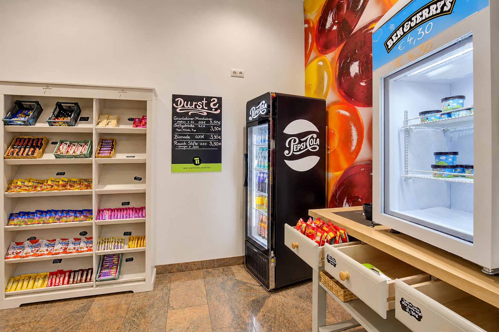 A snack bar with shelves stocked with various packaged snacks, a Pepsi Cola vending machine, and a refrigerator with a glass door.