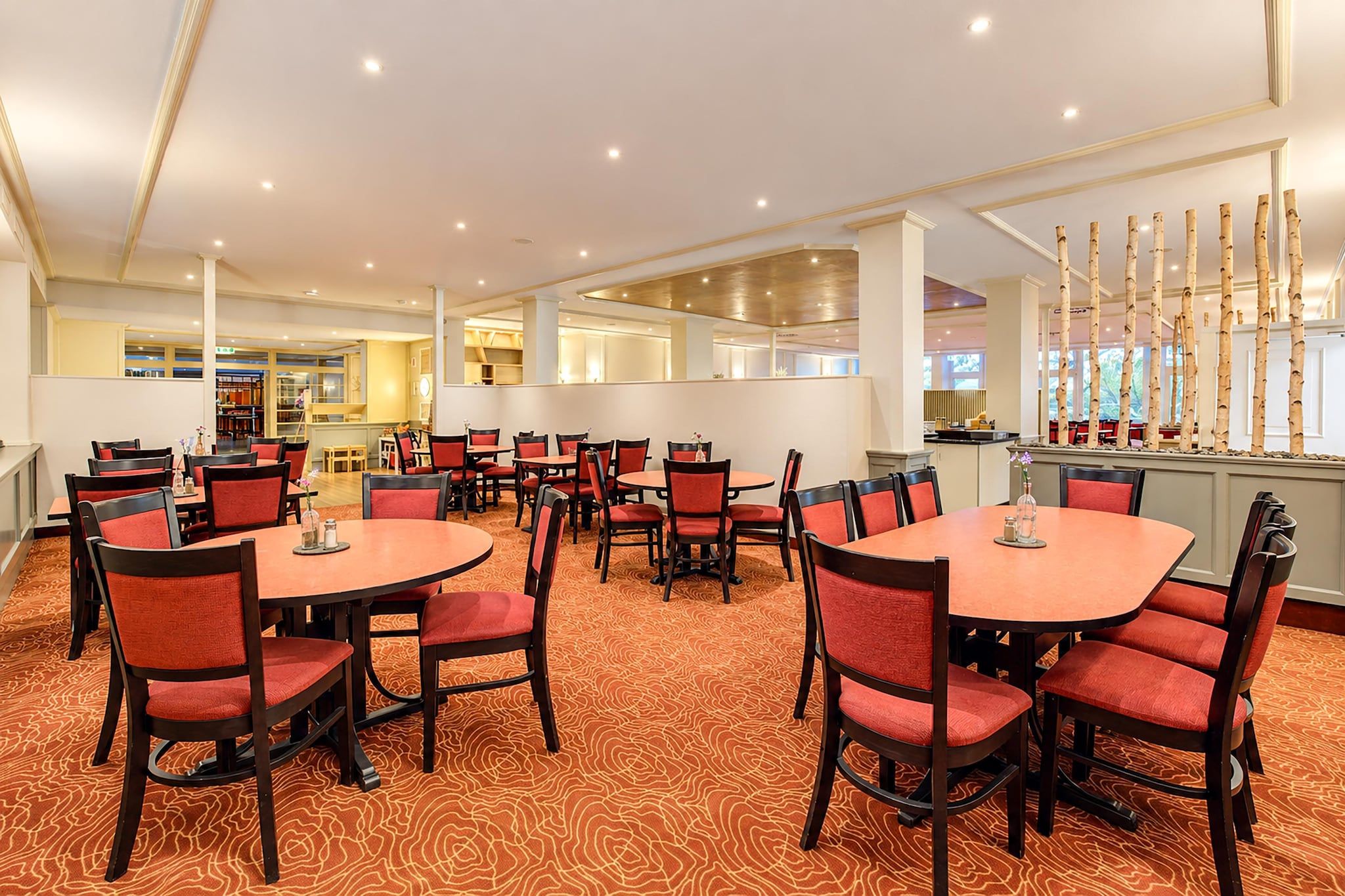 A hotel dining area with round tables and red chairs, a patterned carpet, and a view of the kitchen in the background.