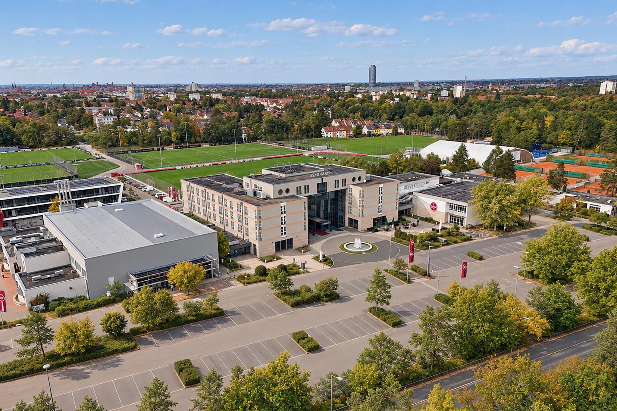 A multi-story building with a parking lot, surrounded by greenery and a sports field.