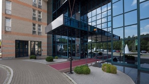 Un edificio moderno con facciata in vetro, sezione inferiore in mattoni e area d'ingresso pavimentata con tappeto rosso e piccoli arbusti.
