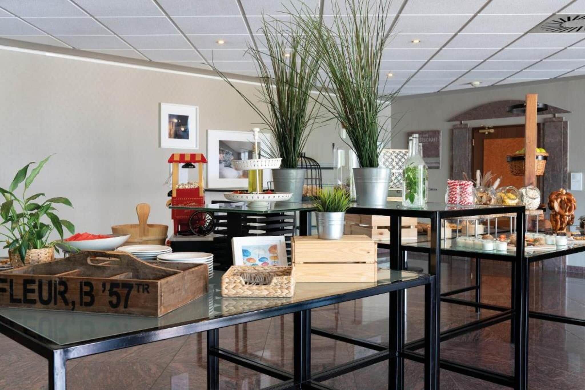 Un interior moderno con una mesa de vidrio que exhibe varios objetos, incluyendo un caja de madera con la etiqueta 'FLEUR, B '57 TR', una máquina de palomitas y plantas decorativas.