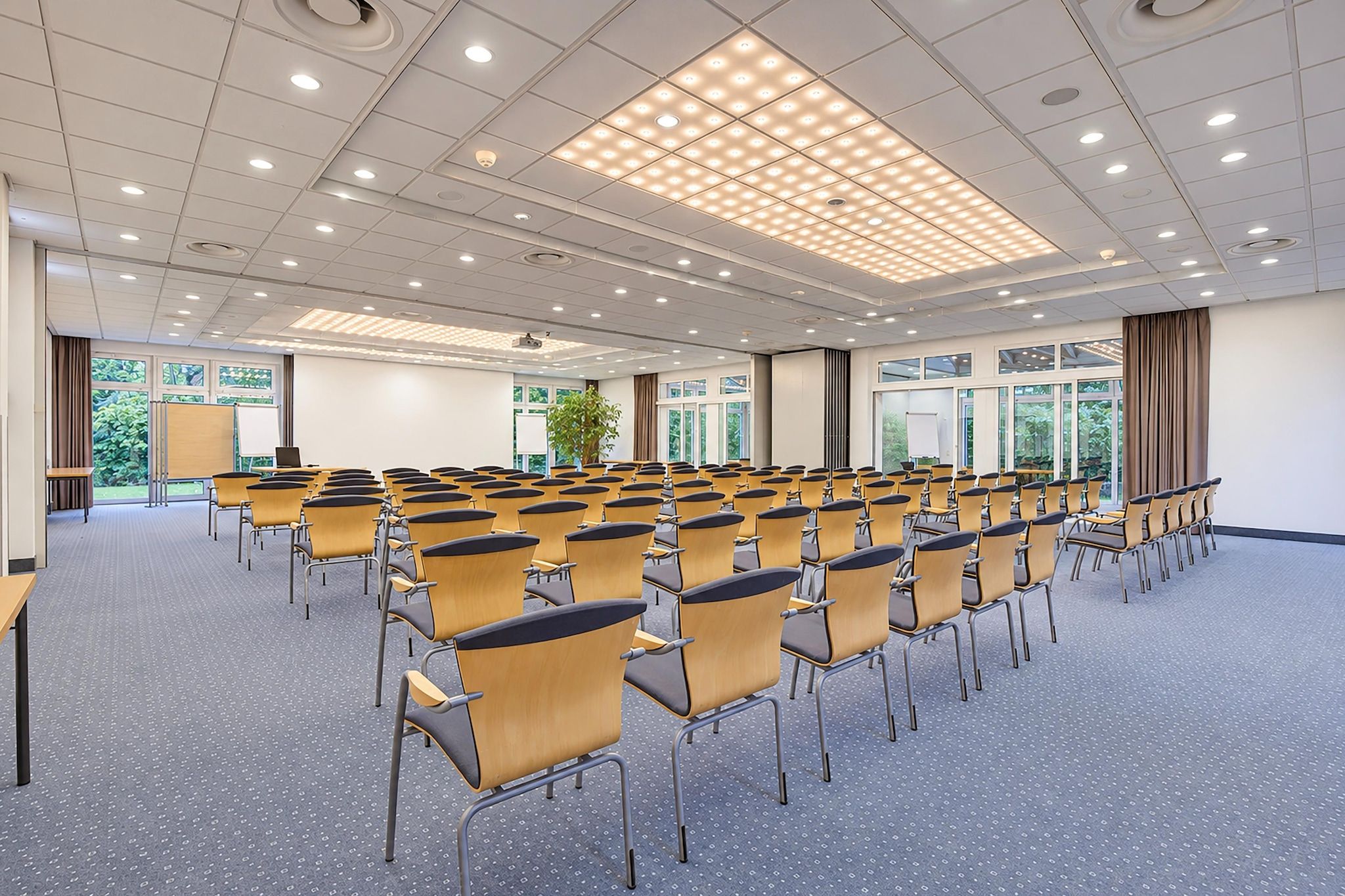 Ein Konferenzraum mit Reihen von Stühlen, einer Projektionsleinwand und einem großen Fenster mit Blick auf Grünflächen.