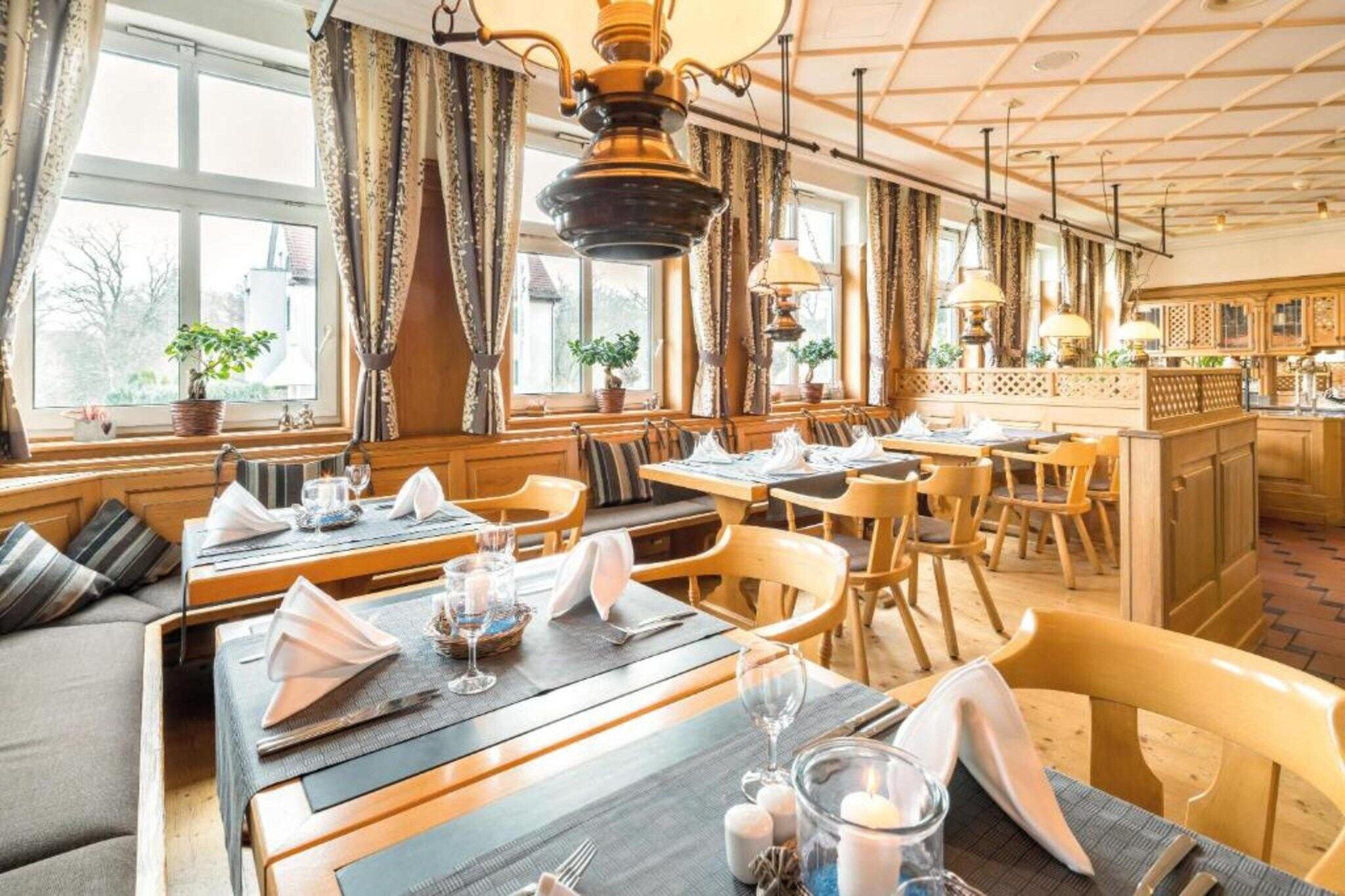 Ein Restaurant mit Holztischen und -stühlen, gedeckten Tischen mit weißen Servietten und Gläsern sowie Blick ins Freie durch große Fenster.
