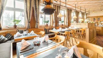 Ein Restaurant mit Holztischen und -stühlen, gedeckten Tischen mit weißen Servietten und Gläsern sowie Blick ins Freie durch große Fenster.