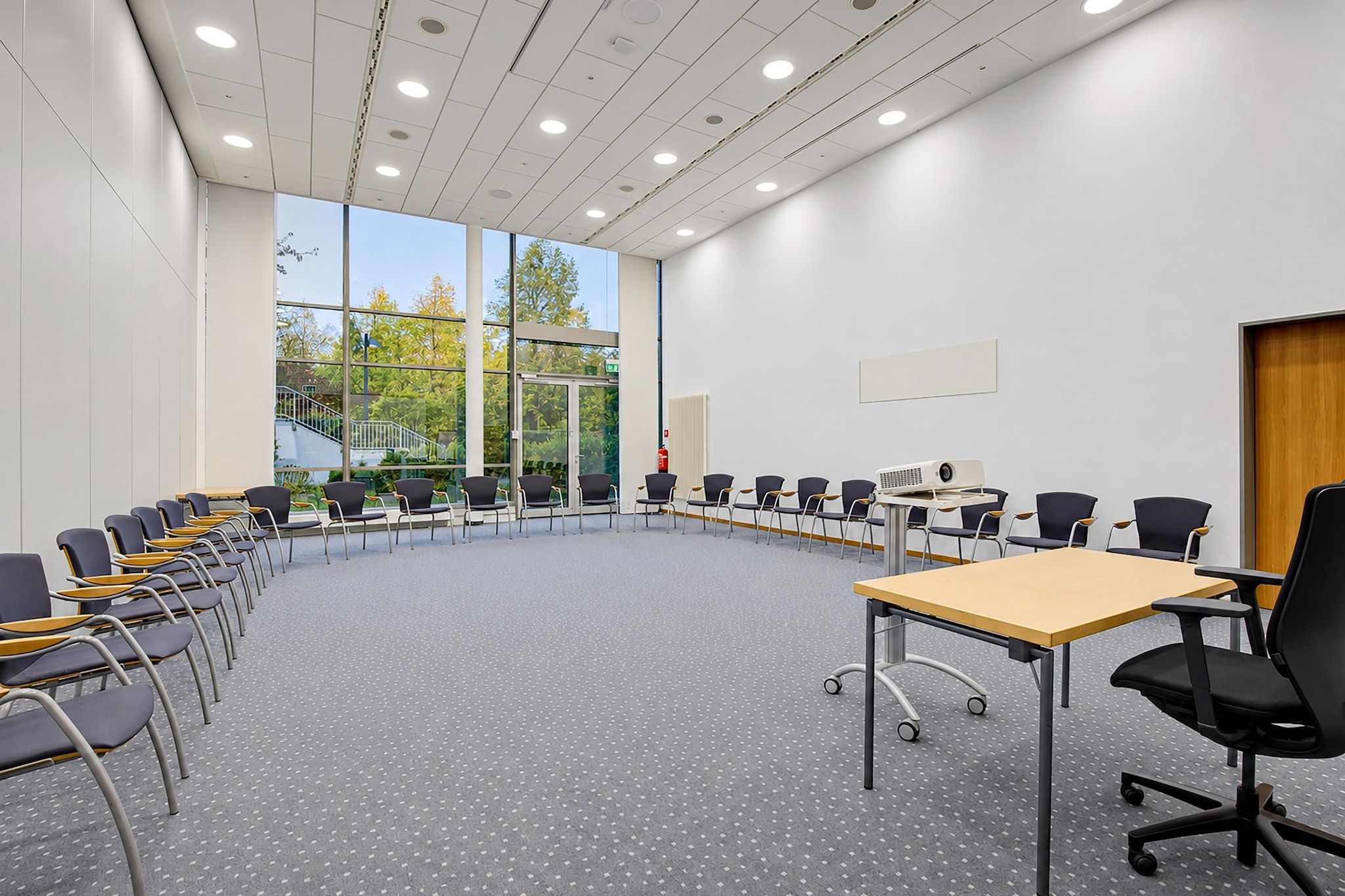 A conference room with a large oval table, multiple chairs, a projector, and large windows offering a view of trees and a building outside.