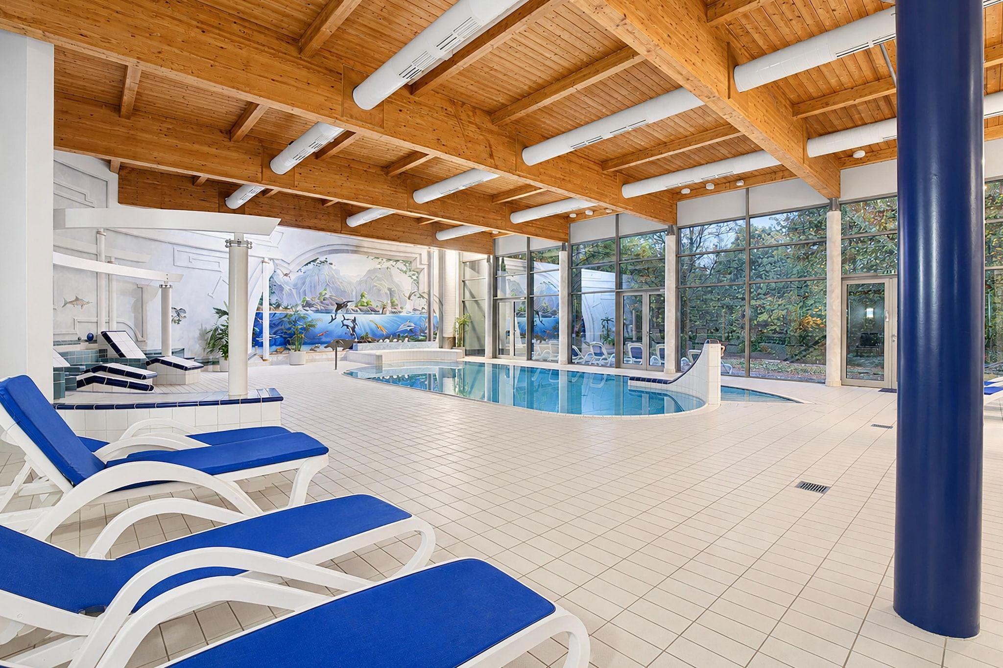 An indoor swimming pool with blue lounge chairs, a mural, and a view of greenery outside.