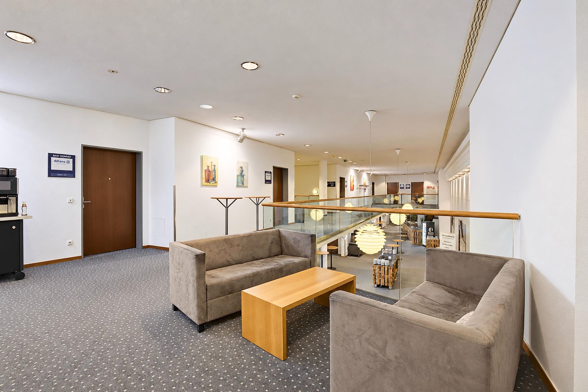 Un vestíbulo de hotel con un mostrador de recepción, una zona de estar con sofás y una mesa de centro, una escalera con barandillas de vidrio y obras de arte en las paredes.