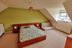 A bedroom with a sloped ceiling, a bed with floral bedding, a bedside lamp, and a window with a view. - Pebble House (Castlerock)