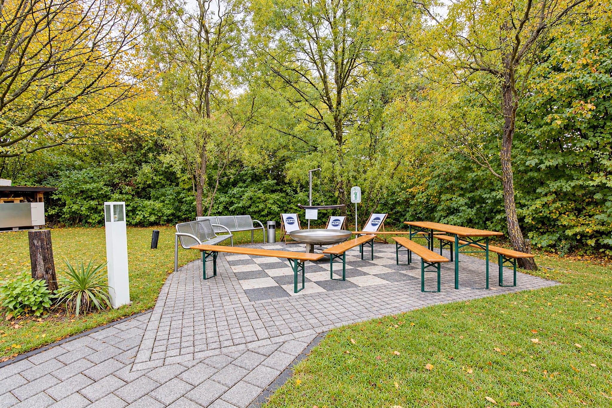 An outdoor seating area with benches and tables, surrounded by trees and a paved walkway.