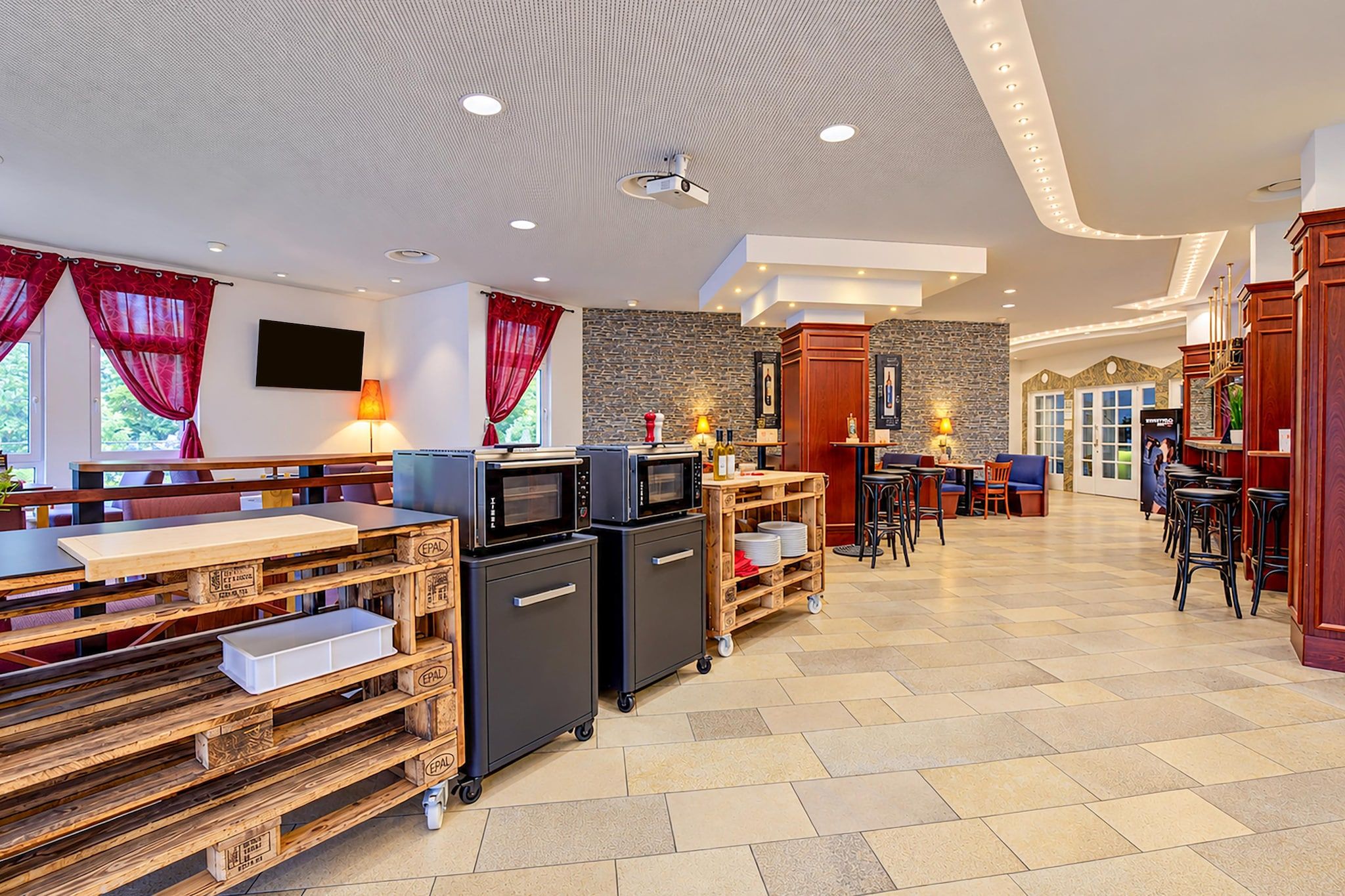 En rummelig hotellobby med et moderne design, der indeholder et siddeområde med borde og stole, et barområde og et køkken med hårde hvidevarer.