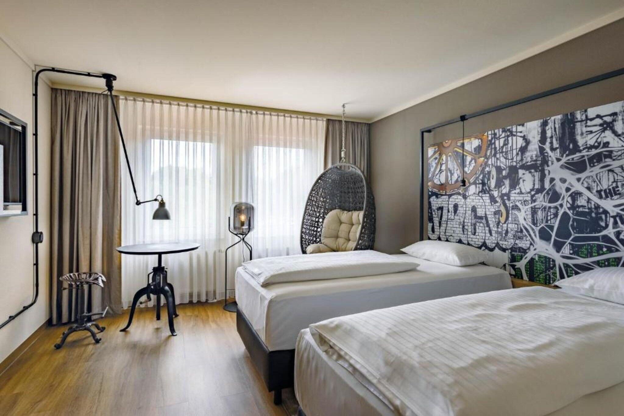 Ein Hotelzimmer mit zwei Betten, einem Hängekorbsessel, einem kleinen Tisch und einem großen, abstrakten Gemälde an der Wand.