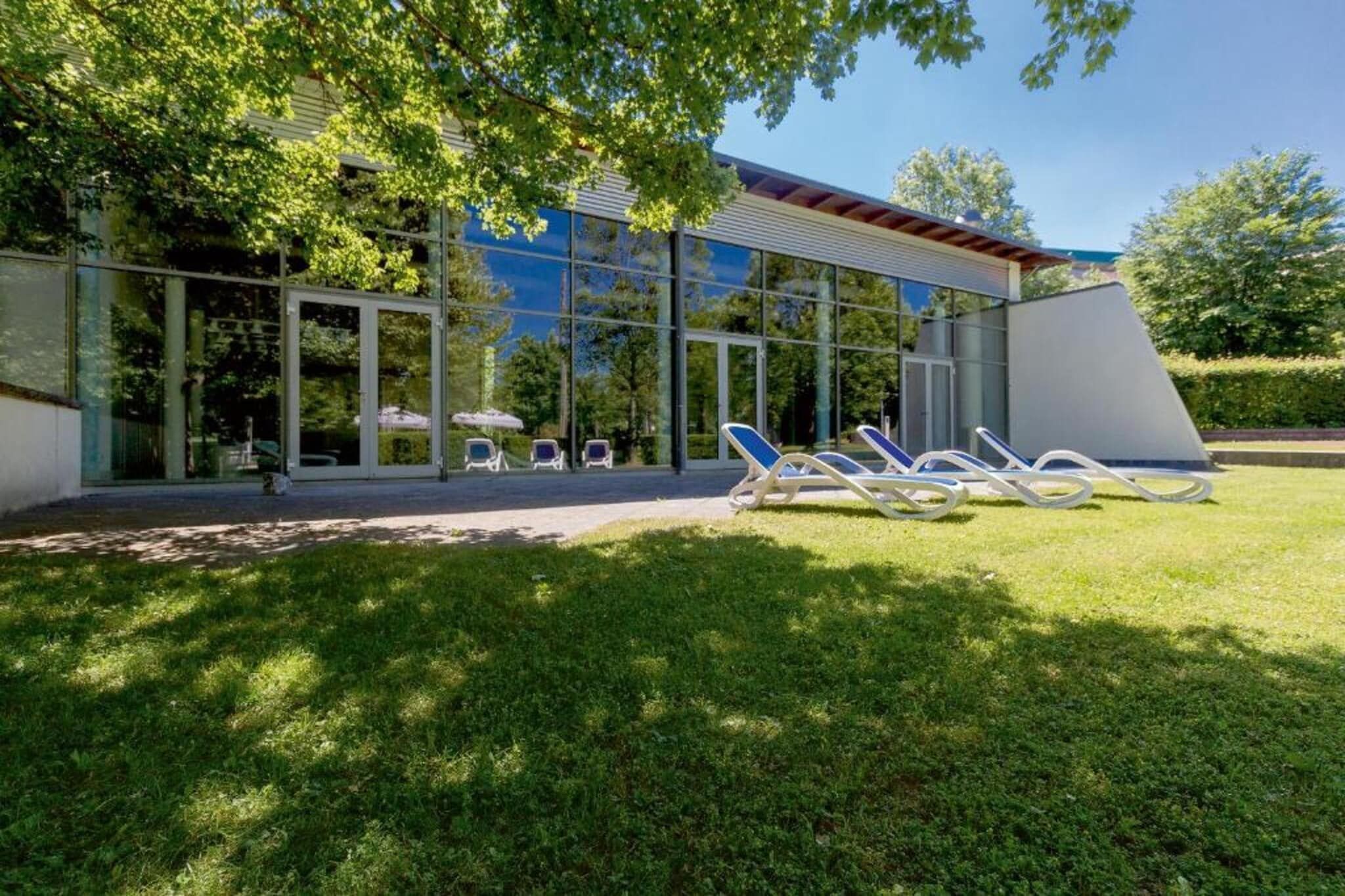 Ein modernes Gebäude mit großen Glasfenstern, einer überdachten Außenbereich und mehreren Liegestühlen auf einer Rasenfläche.