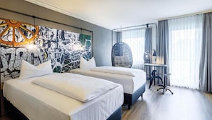 Una camera d'albergo con due letti, un quadro astratto di grandi dimensioni appeso al muro, una poltrona sospesa e una scrivania con una lampada.