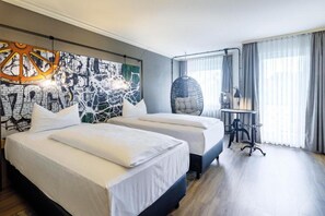 Ein Hotelzimmer mit zwei Betten, einem großen abstrakten Gemälde an der Wand, einem Hängesessel und einem Schreibtisch mit Lampe.
