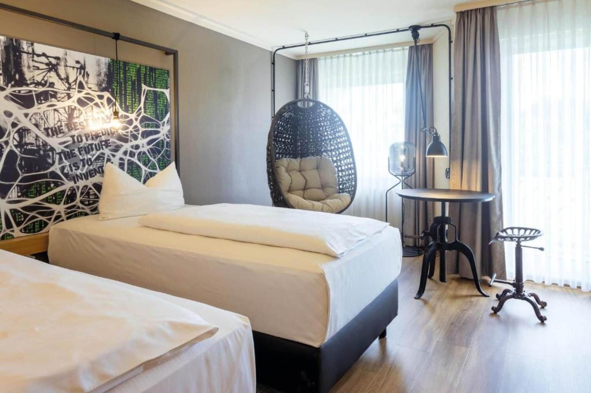 A hotel room with a bed, a hanging chair, a desk with a lamp, and a wall with abstract art.