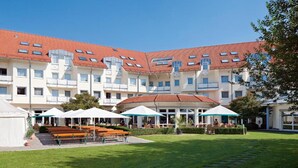 Un hotel a più piani con un tetto in tegole rosse, una zona salotto esterna con tavoli e sedie, e un prato curato.