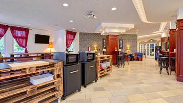 A spacious hotel lobby with a modern design, featuring a seating area with tables and chairs, a bar area, and a kitchen with appliances.