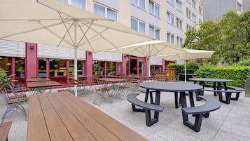 An outdoor seating area with tables and chairs, large umbrellas, and a building with windows in the background.