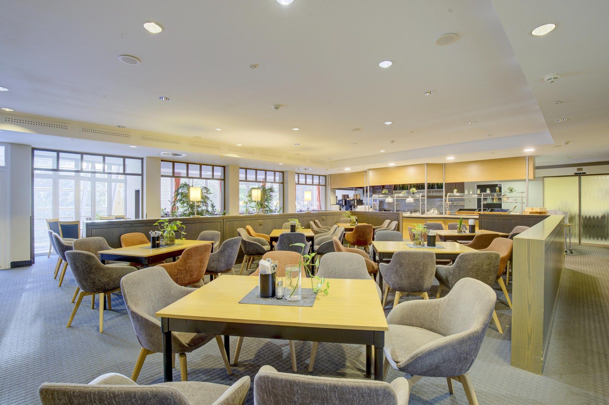 A modern dining area with multiple tables and chairs, a kitchen in the background, and large windows allowing natural light.