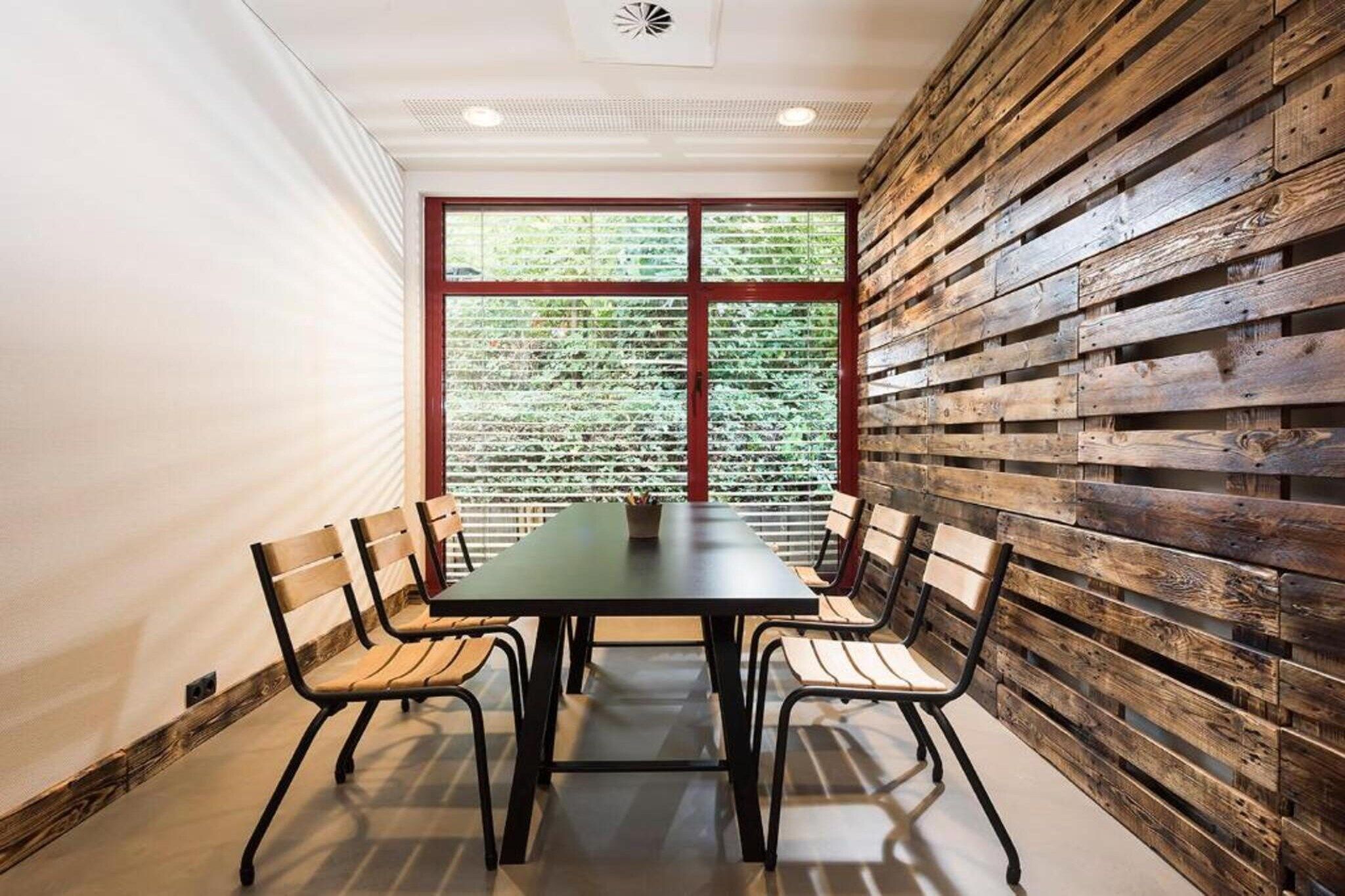 A conference room with a long table and four chairs, a window with blinds, and a wooden wall panel.