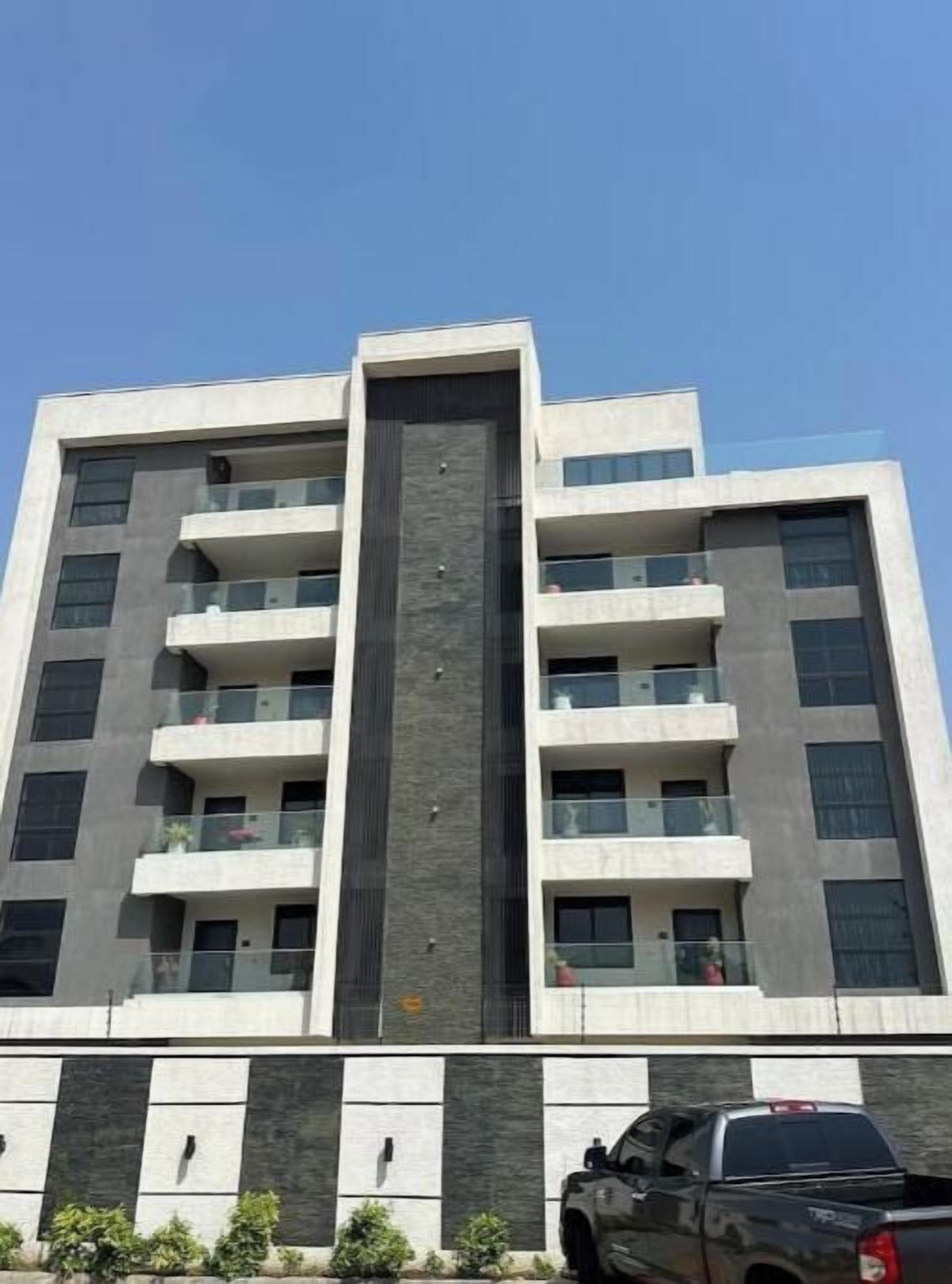 Un edificio de varios pisos con balcones, un elemento vertical central y un área de estacionamiento con un camión.