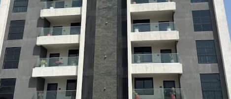 Un edificio de varios pisos con balcones, un elemento vertical central y un área de estacionamiento con un camión.