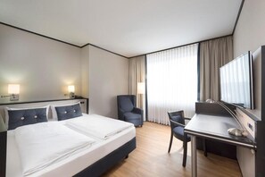 Quarto de hotel com cama, escrivaninha, cadeira, televisão e cortinas.