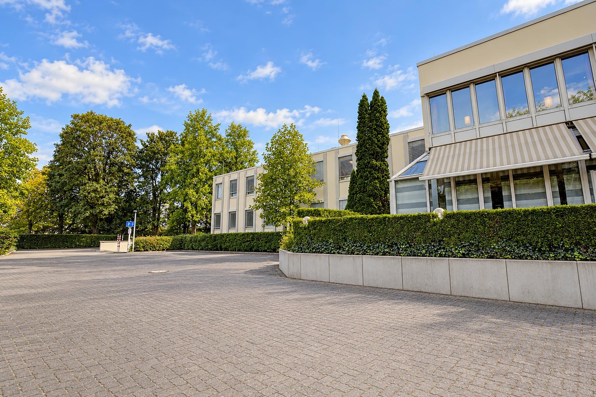 Ein gepflasterter Innenhof mit einem modernen Gebäude, einem Parkschild und ordentlich geschnittenen Hecken.