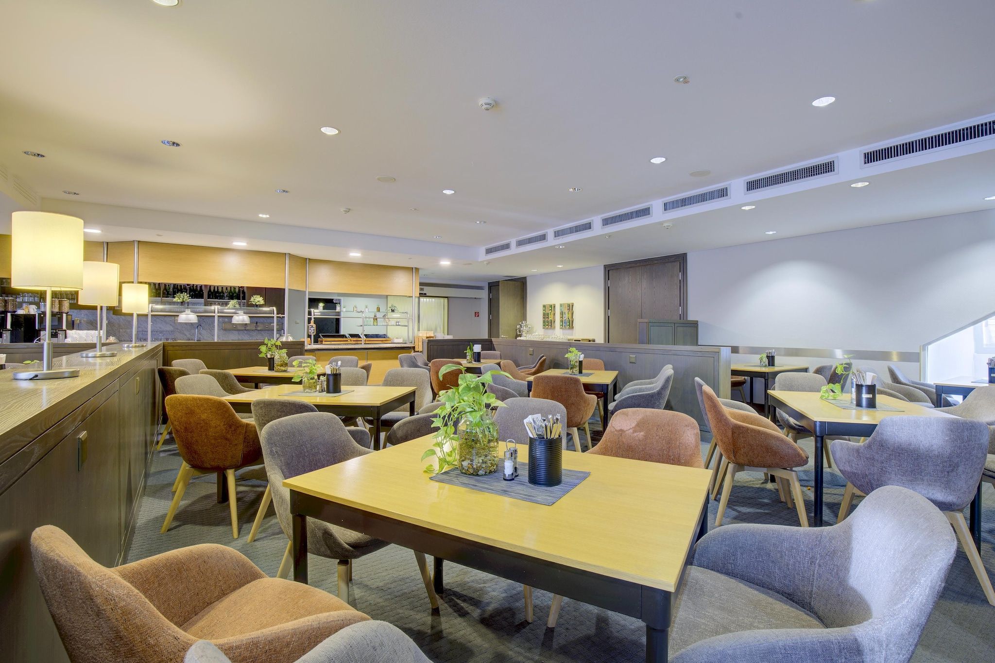 A modern hotel dining area with multiple tables and chairs, a bar area, and a kitchen in the background.