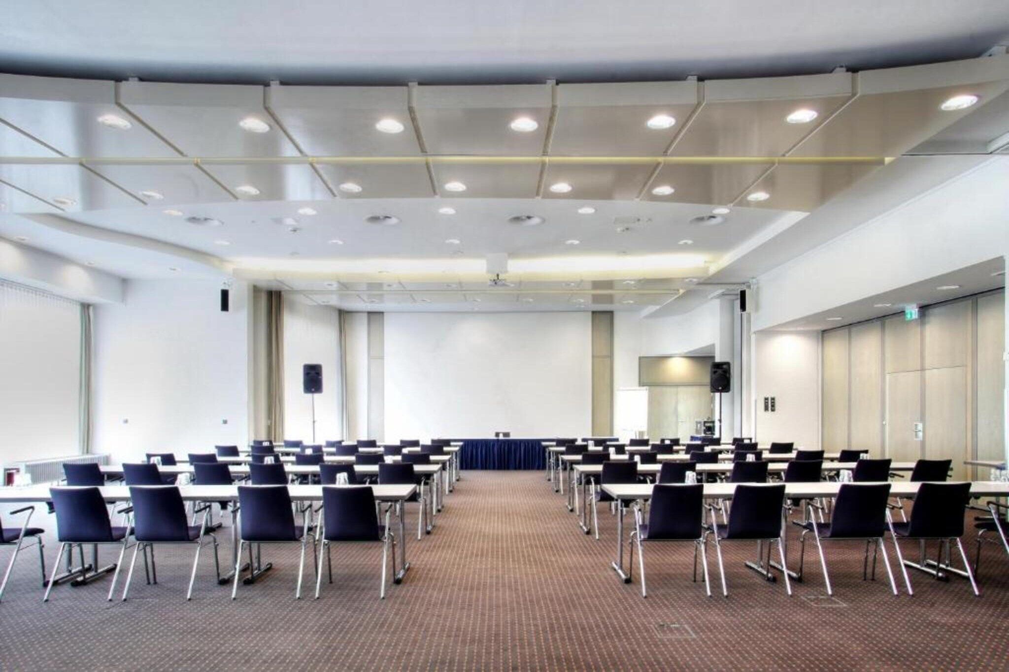 Konferenssisali, jossa on riveissä pöytiä ja tuoleja, puhujapönttö ja matolla peitetty lattia.