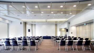 Konferenssisali, jossa on riveissä pöytiä ja tuoleja, puhujapönttö ja matolla peitetty lattia.