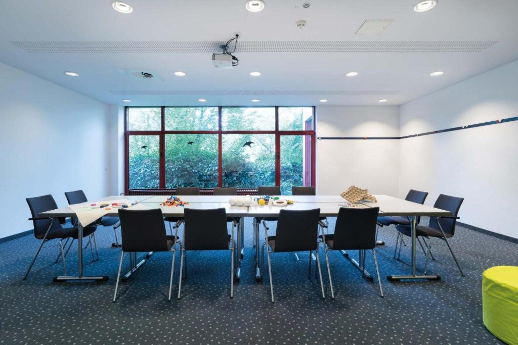 A conference room with a long table, chairs, a projector, and a view of trees through large windows.