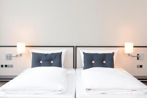 A hotel room with two beds, each with white bedding and dark pillows, and wall-mounted lamps.