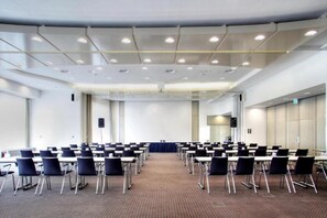Un salón de conferencias con filas de mesas y sillas, un atril y piso alfombrado.