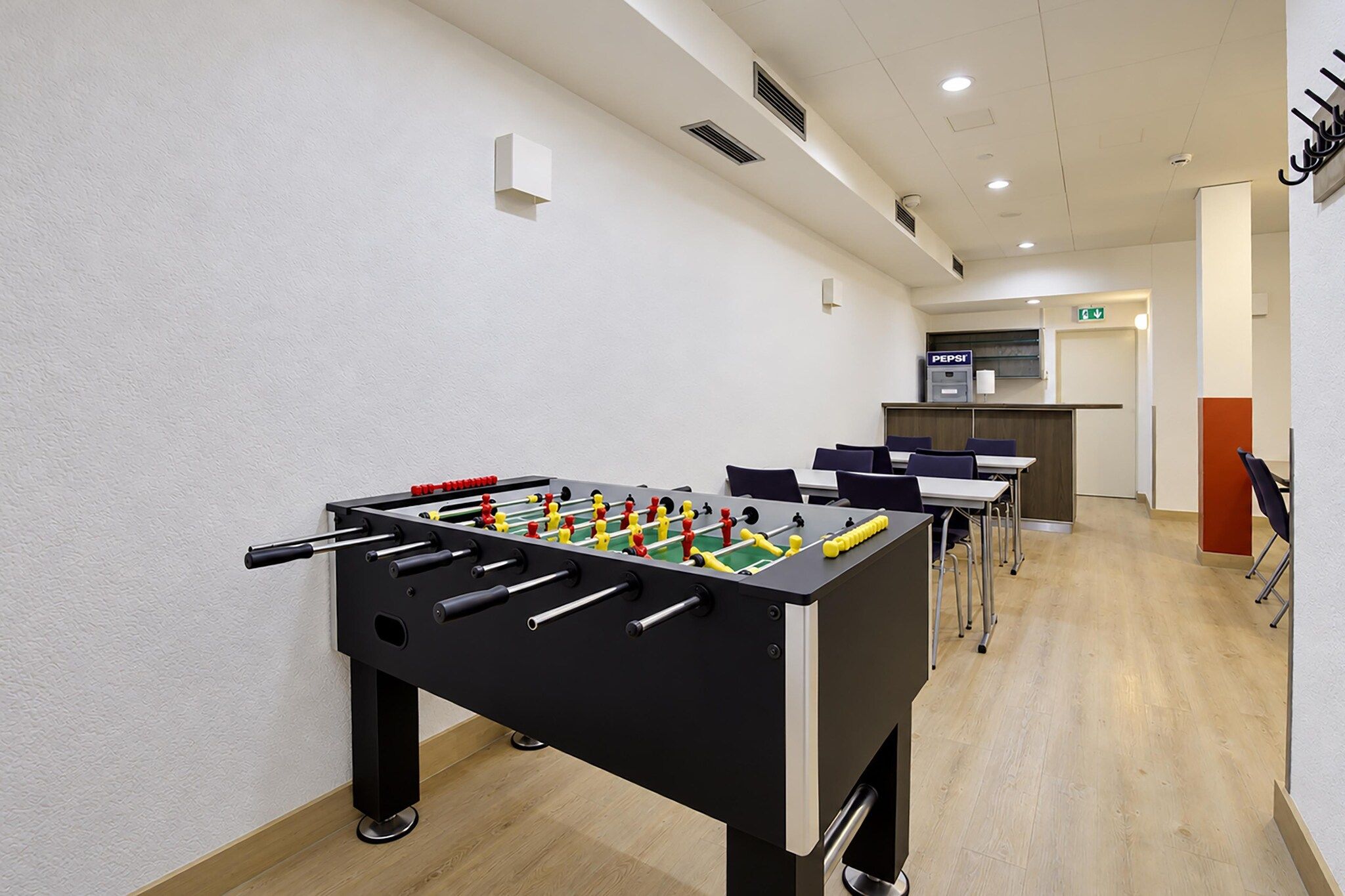 Une pièce équipée d’un baby-foot, d’un coin cuisine et d’un espace détente avec des tables et des chaises.