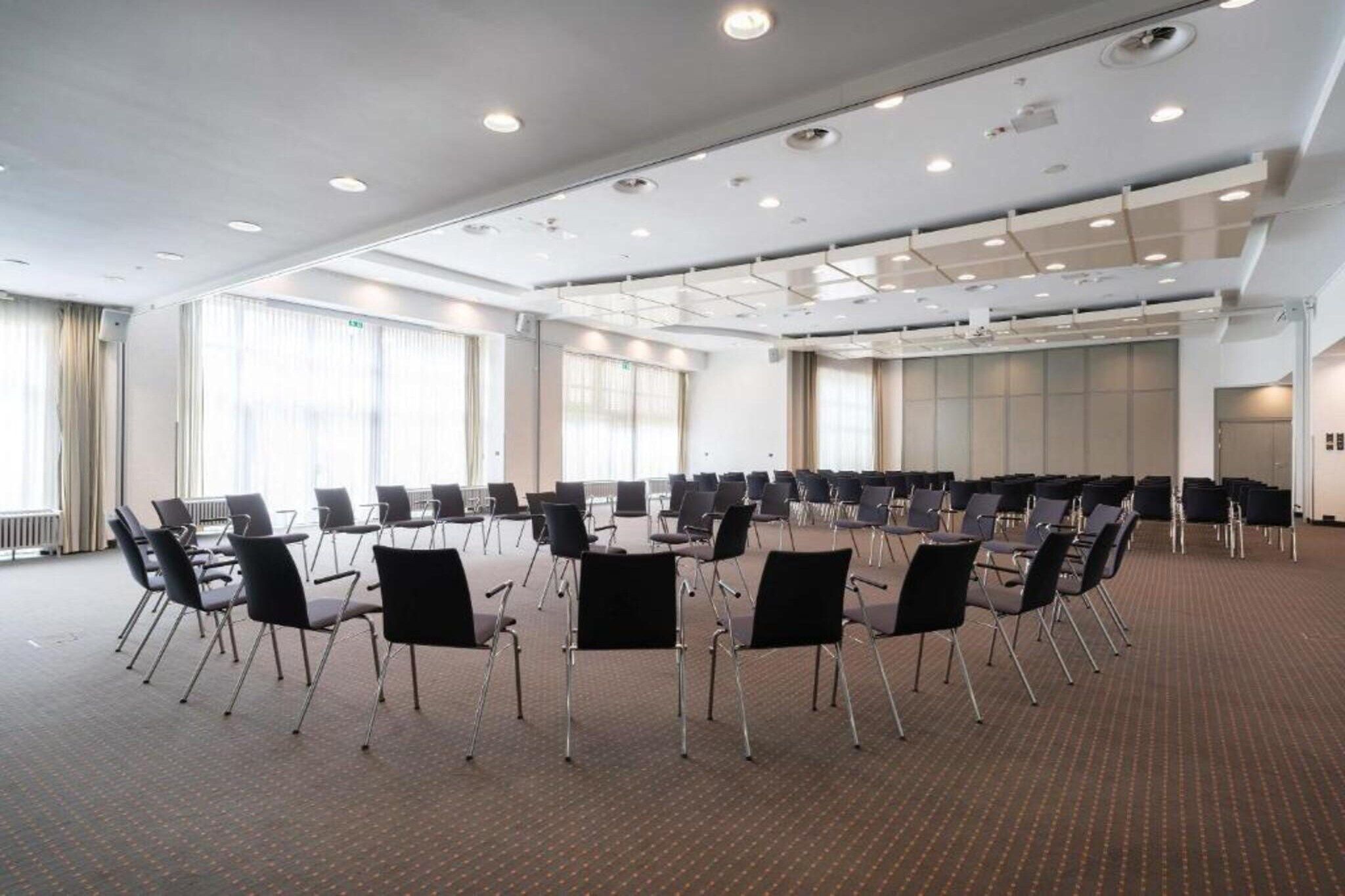 Konferenssihuone, jossa on riveissä tuoleja, suuret ikkunat ja matolla peitetty lattia.