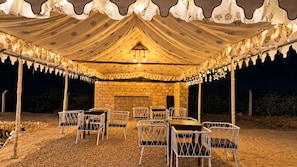 Un espace de restauration en plein air, avec un mur en pierre en arrière-plan, des chaises en métal blanc et une tente recouverte d’un tissu à motifs.