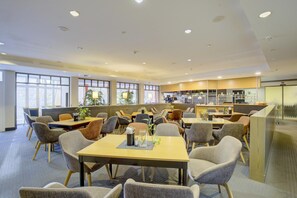 Un comedor moderno con varias mesas y sillas, una cocina al fondo y grandes ventanas que dejan entrar luz natural.