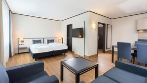 Quarto de hotel moderno com cama de casal, mesa pequena, duas cadeiras, escrivaninha e TV fixada na parede.