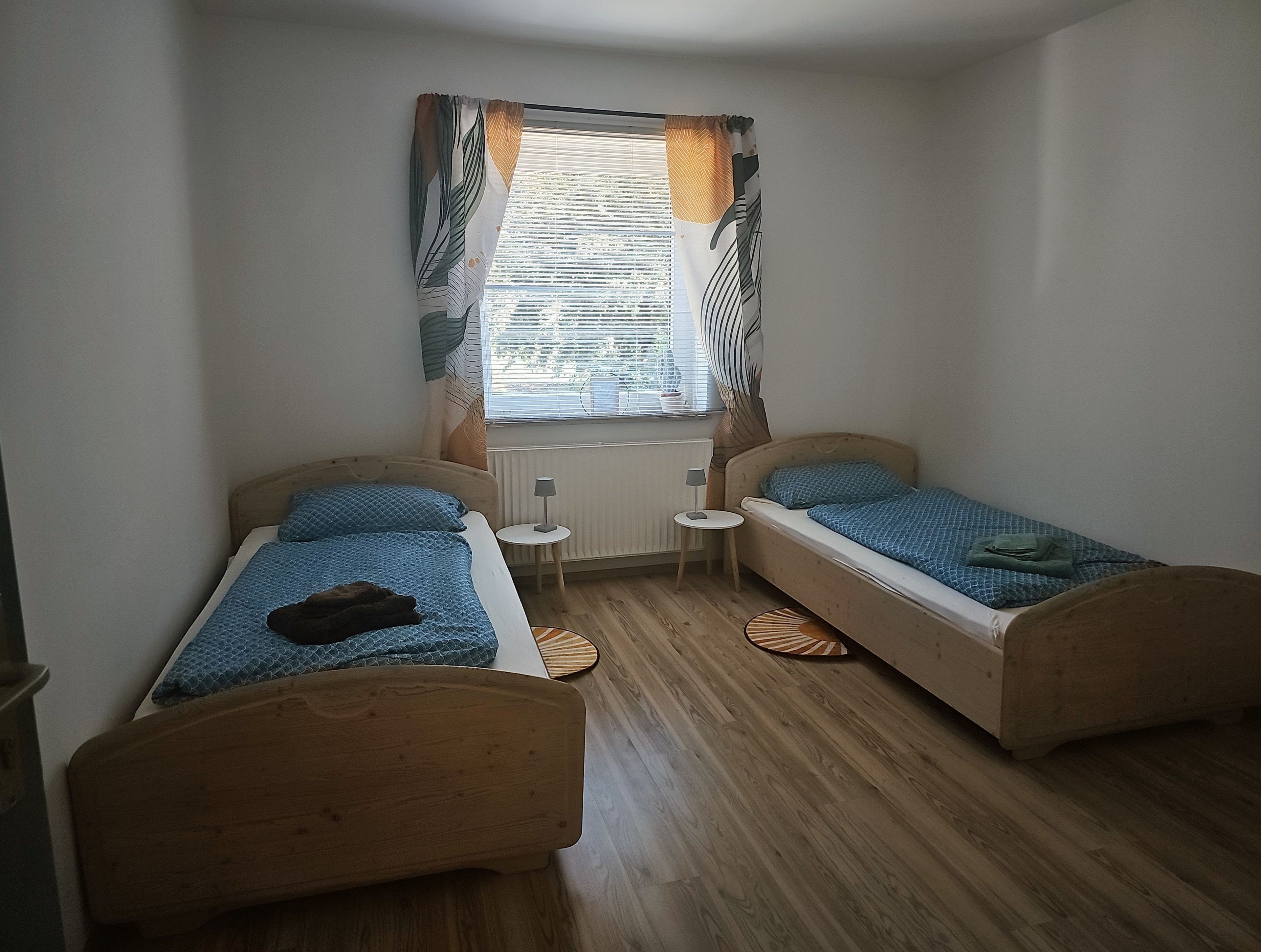 Un petit logement composé d’une seule pièce, comprenant deux lits en bois, une fenêtre avec des rideaux, un radiateur et une petite table.