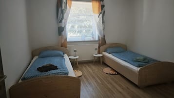 Un petit logement composé d’une seule pièce, comprenant deux lits en bois, une fenêtre avec des rideaux, un radiateur et une petite table.
