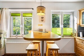 Una cocina moderna con una mesa de madera, asientos tipo banco, una lámpara colgante y ventanas con persianas verdes.