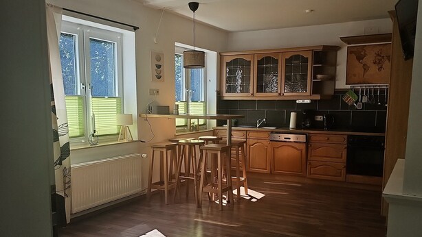 A kitchen with wooden cabinets, a dining area with stools, and a view of the outdoors through a window. - Apartment 1 – Schrappmühle with Terrace, Garden, Wi-Fi, and Rural Seclusion (Aerzen)