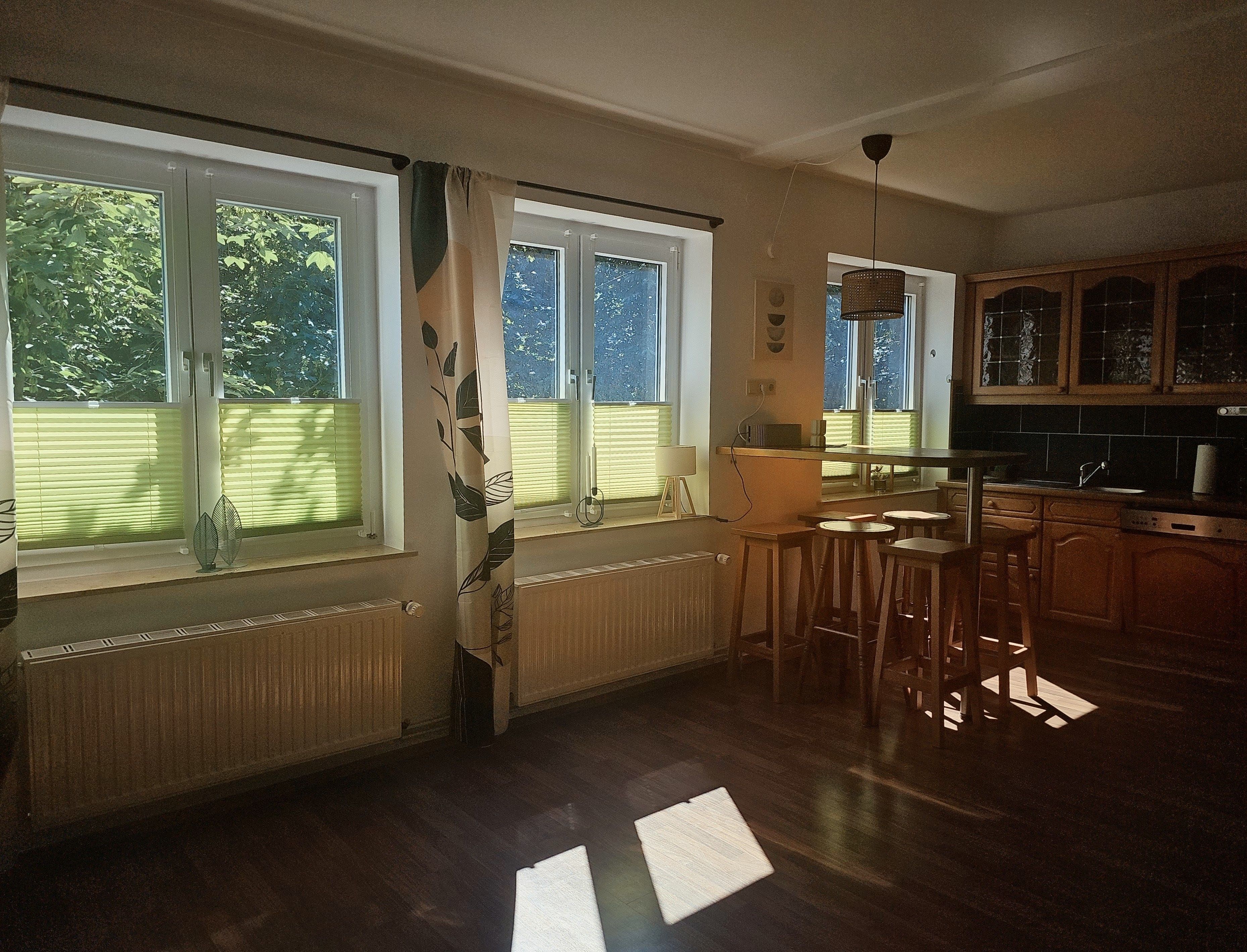 A kitchen with wooden cabinets, a dining area with stools, and large windows with blinds.