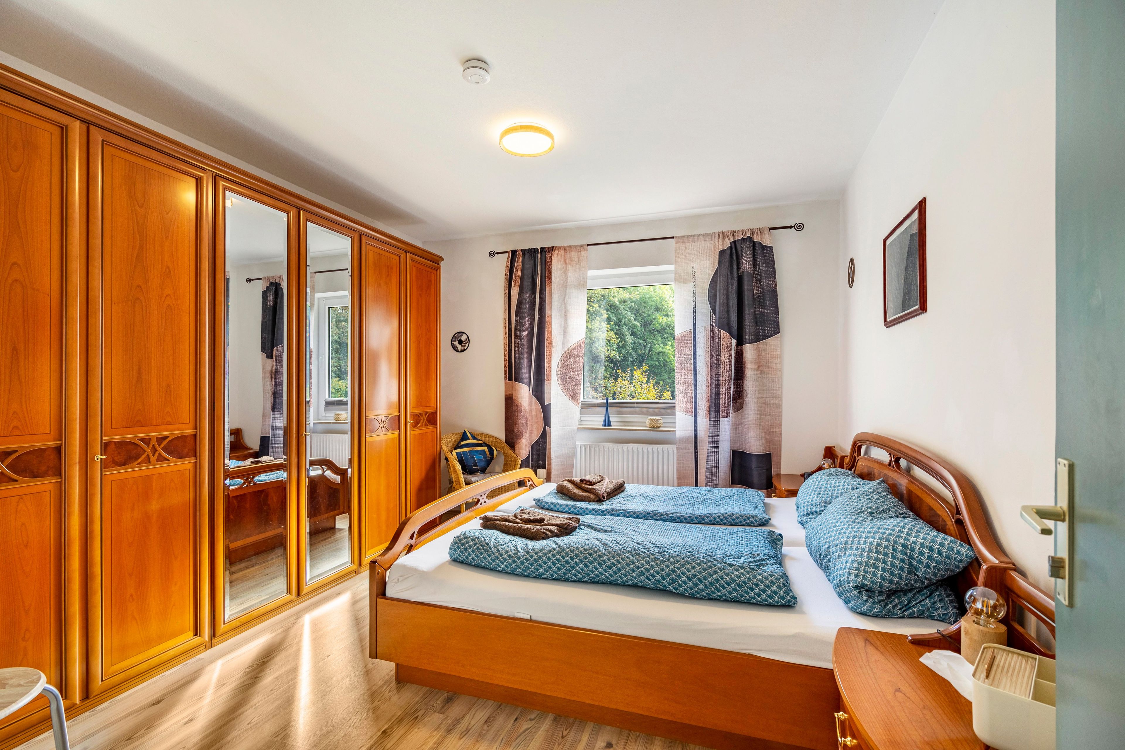 Ein Schlafzimmer mit einem Holzschrank, einem Bett mit gemusterten Betttextilien, einem Fenster mit Vorhängen und einem Nachttisch.