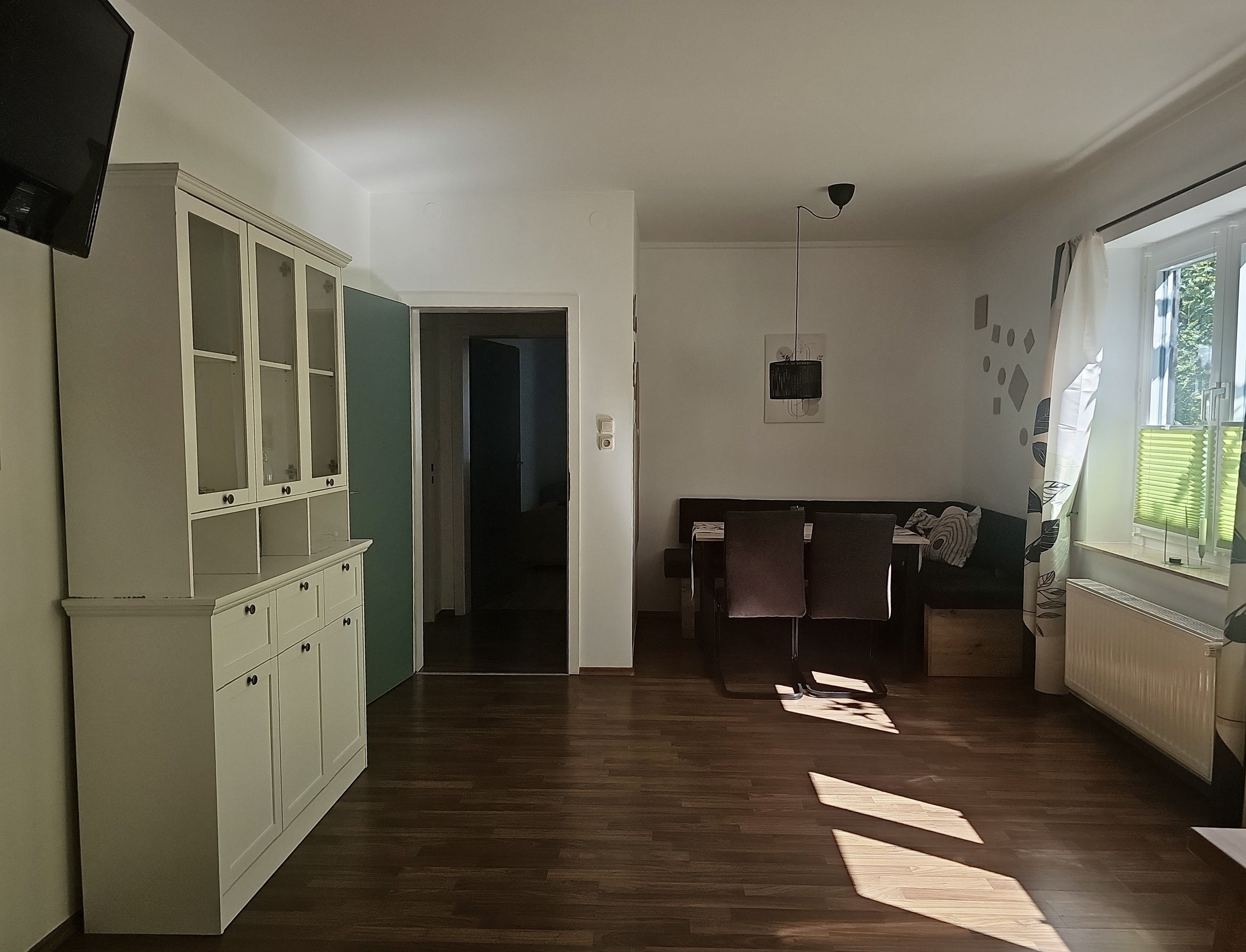 Une pièce avec un plancher en bois, une armoire aux portes vitrées, un lit avec des oreillers, un luminaire suspendu et une fenêtre avec des stores.