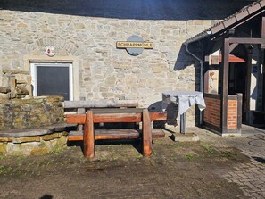 Ein Steinhaus mit einer hölzernen Bank und einem Tisch, einer Tür und einem Schild mit der Aufschrift 'SCHRAPPMÜHLE'.