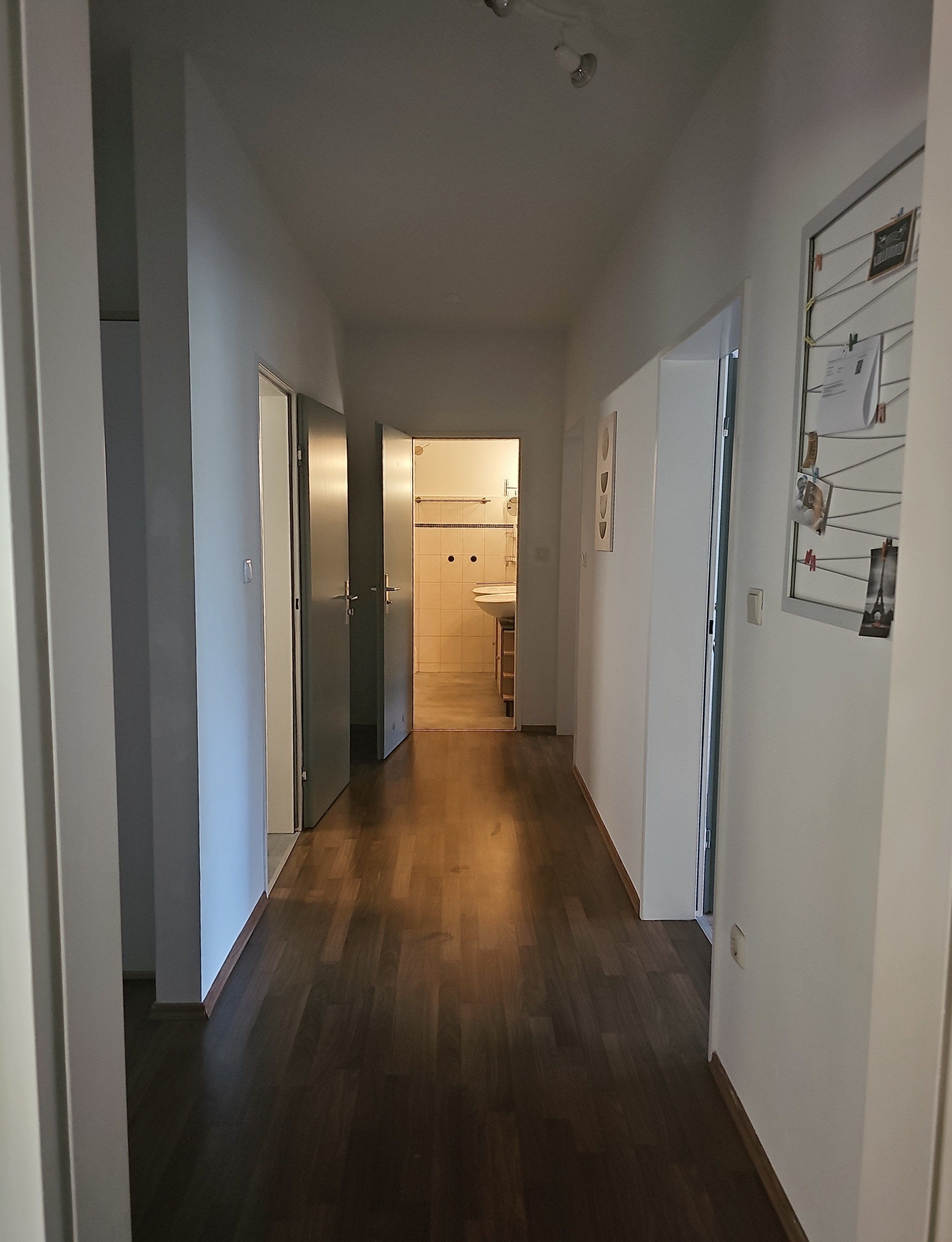 A hallway with wooden flooring, a door on the left, and a wall with a bulletin board on the right.