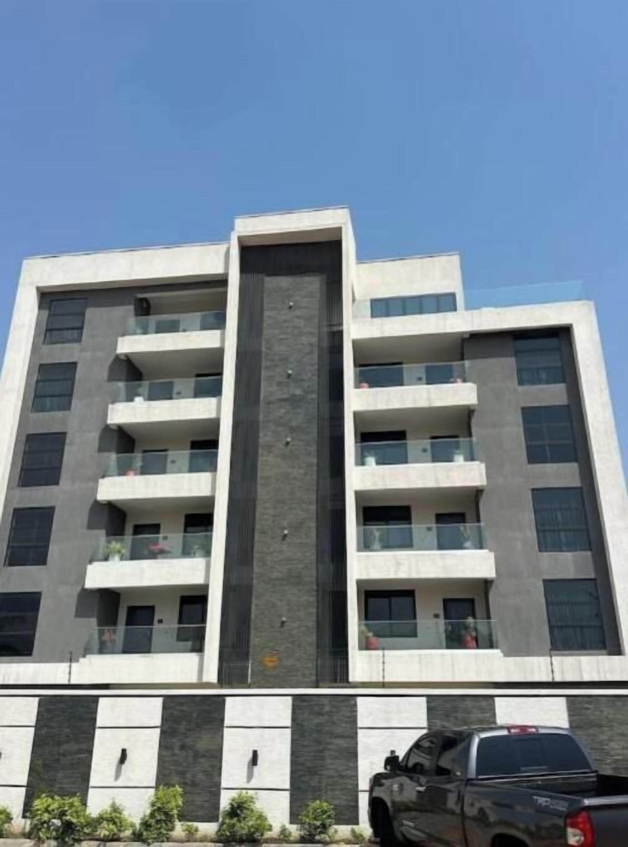 A multi-story building with balconies, a central vertical feature, and a parking area with a truck.